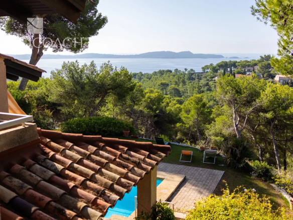 Terraced villa with a red tiled roof and a sunny garden, pool, and sea view beyond trees.