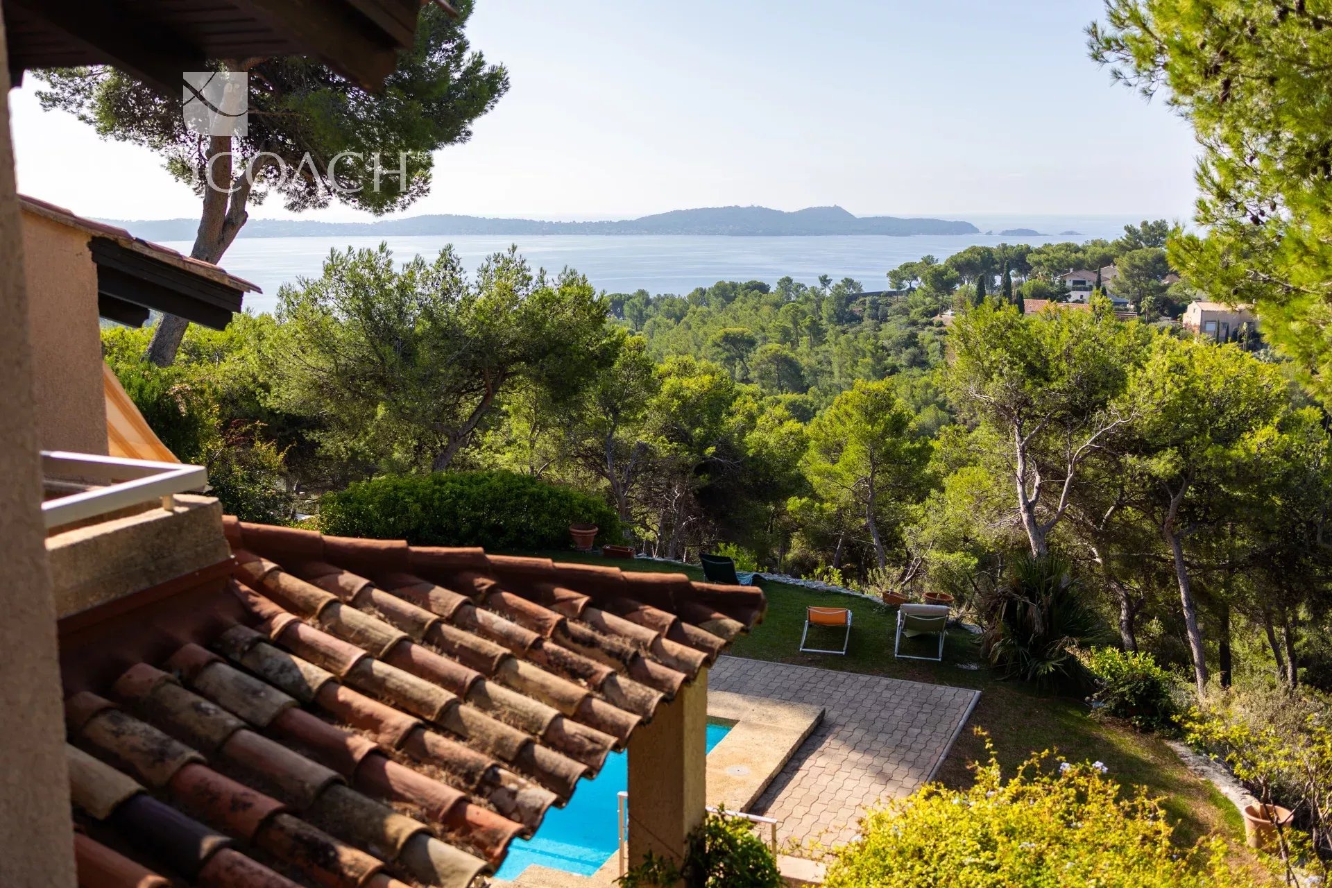 Terraced villa with a red tiled roof and a sunny garden, pool, and sea view beyond trees.