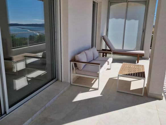Balcony seating area with beige cushions, a lounge chair, a small wooden table, and a view of the blue sea beyond.