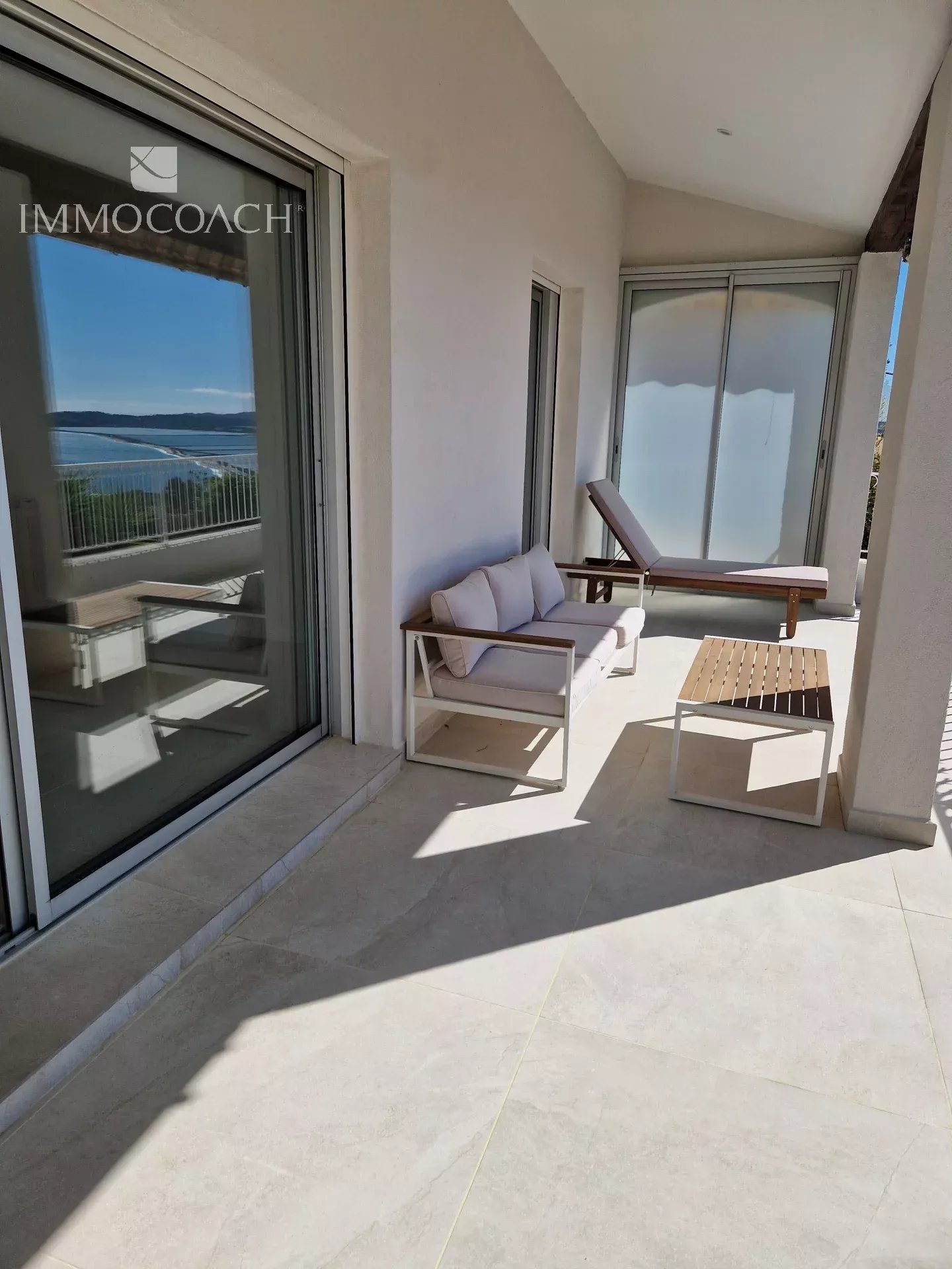 Balcony seating area with beige cushions, a lounge chair, a small wooden table, and a view of the blue sea beyond.