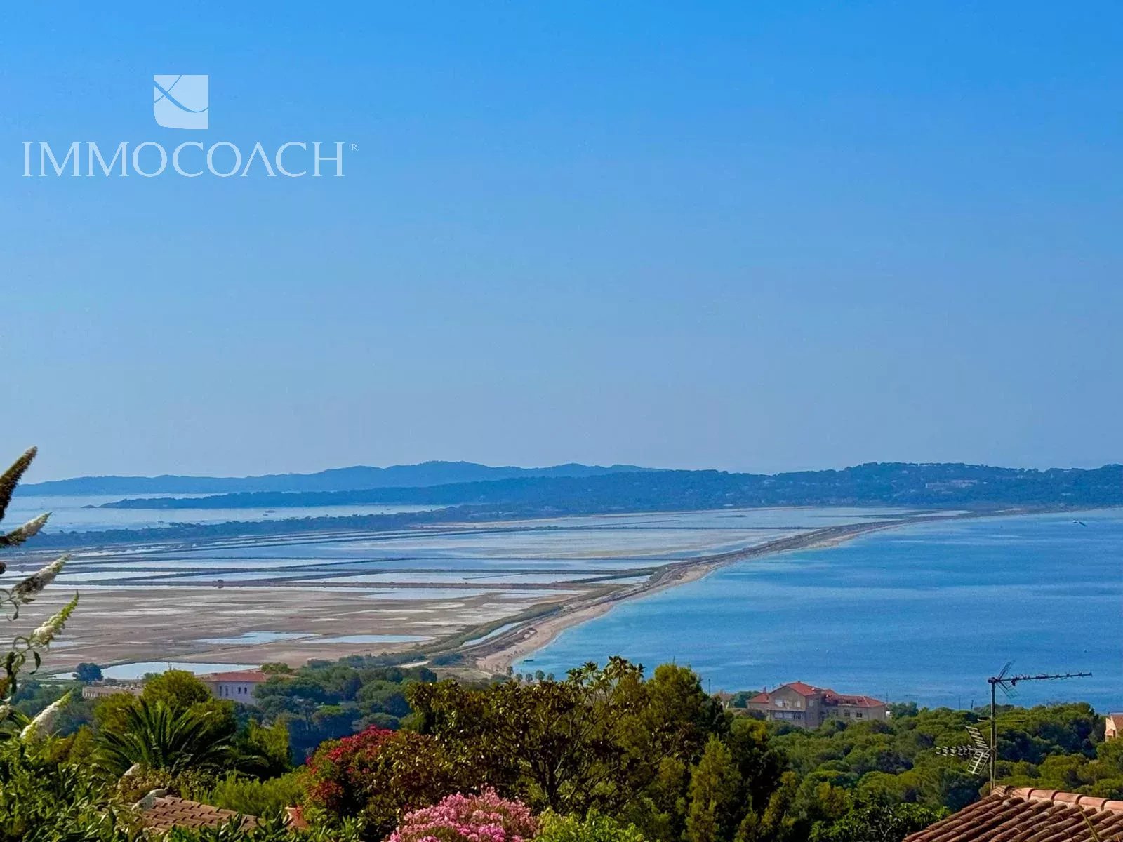 Coastal landscape with blue sea and salt pans along the shore, distant hills, and foreground trees and rooftops.
