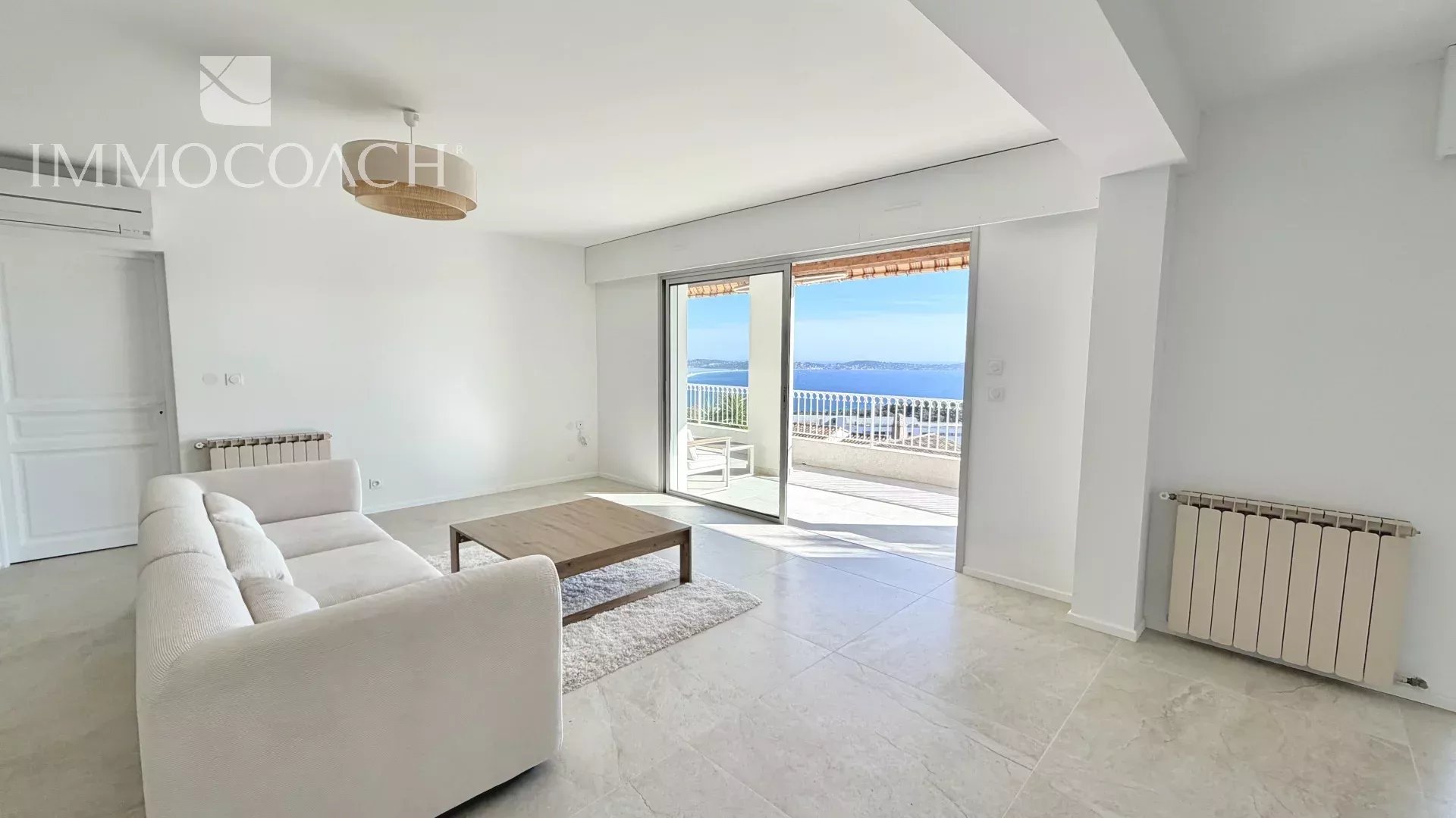 Bright living room with white sofa, wooden coffee table, and a balcony door revealing an ocean view.