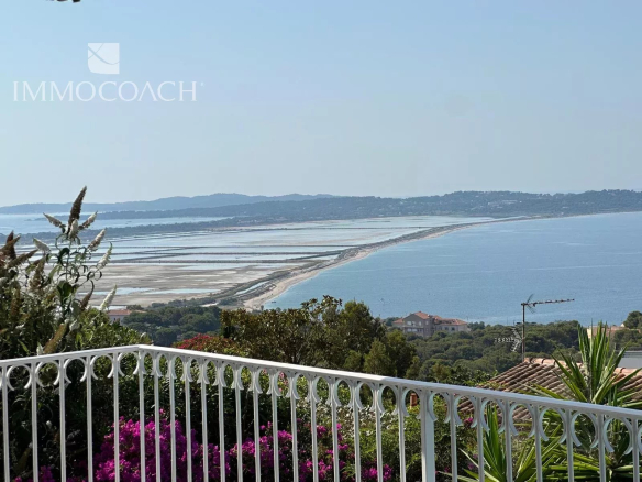 Coastal scene viewed from a terrace with a white decorative railing, pink flowers, and a calm blue bay beyond.