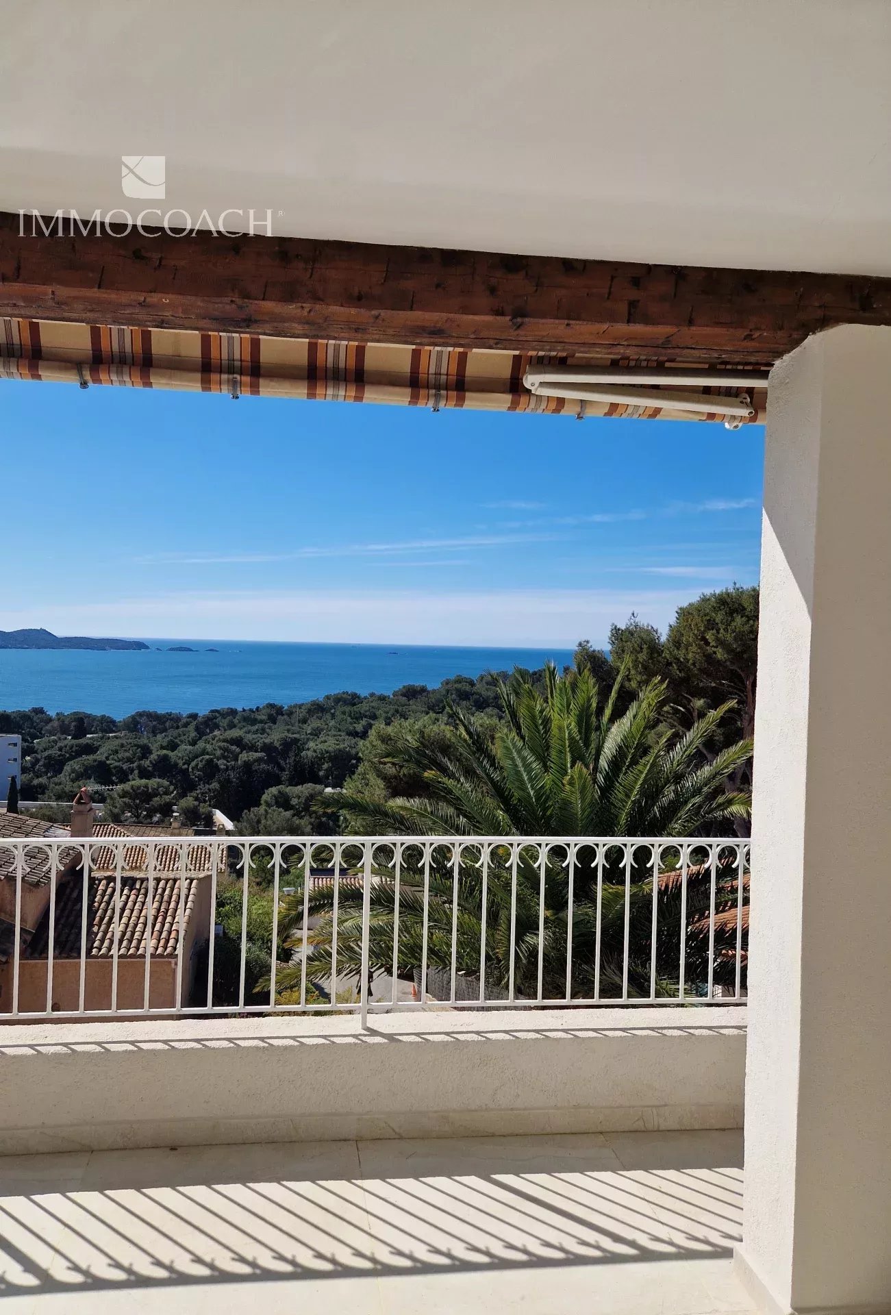 Balcony railing overlooking a blue sea with green trees and a striped awning above.