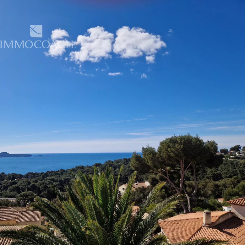 Coastal hillside with red-tiled roofs, palm trees, and a blue sea under a clear sky; tranquil seaside view.