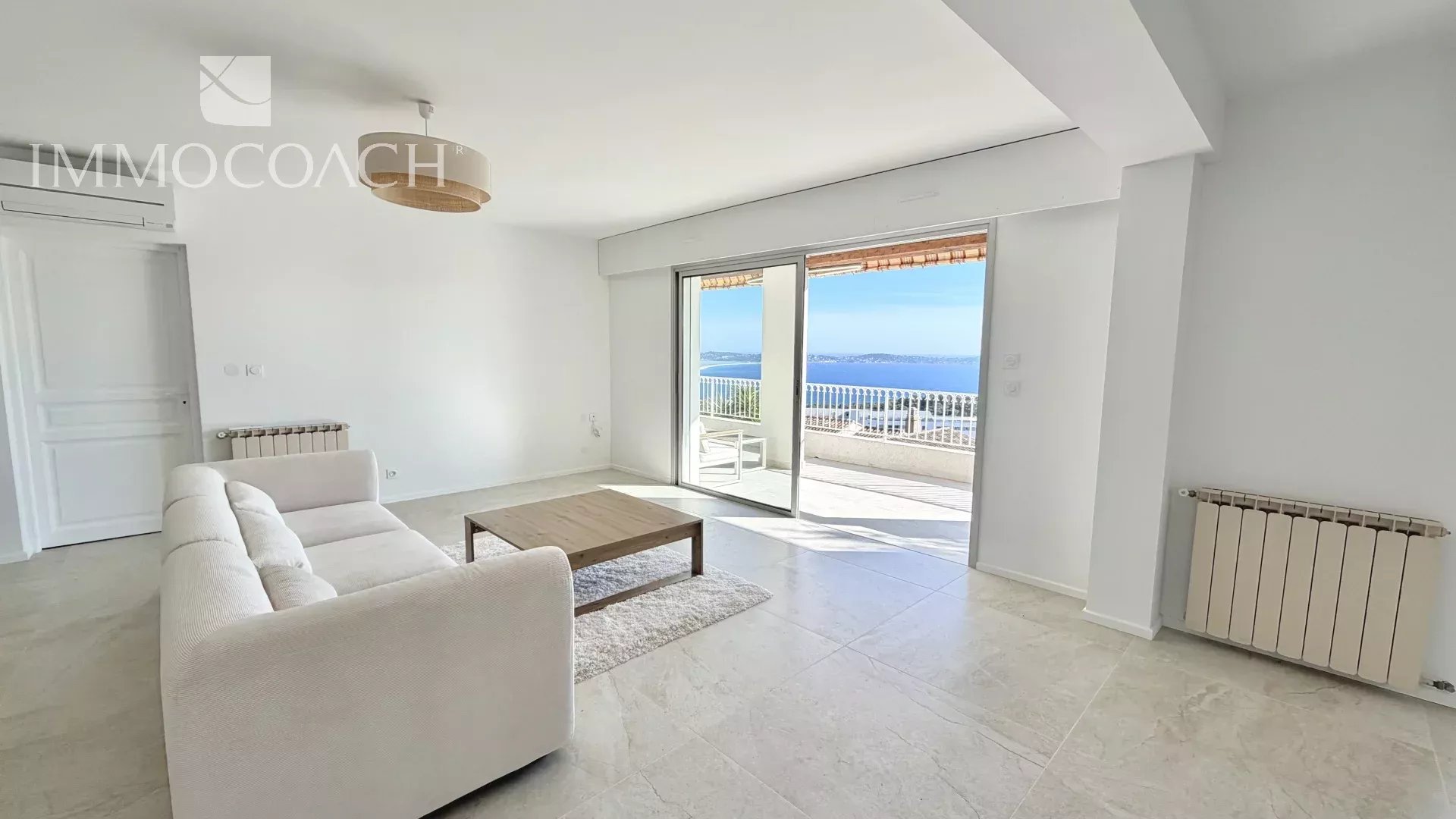 Bright living room with white sofa and wooden coffee table, sliding doors opening to a balcony with ocean view.