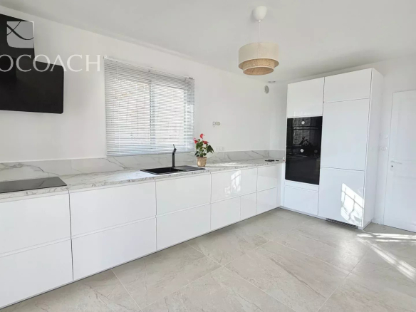 Bright white minimalist kitchen with marble countertops, long handleless cabinets, and a built-in oven cluster.