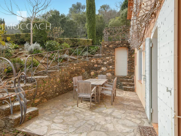 Stone patio with a wooden table and six chairs, surrounded by stone walls and garden, with a door and shutters on the right.