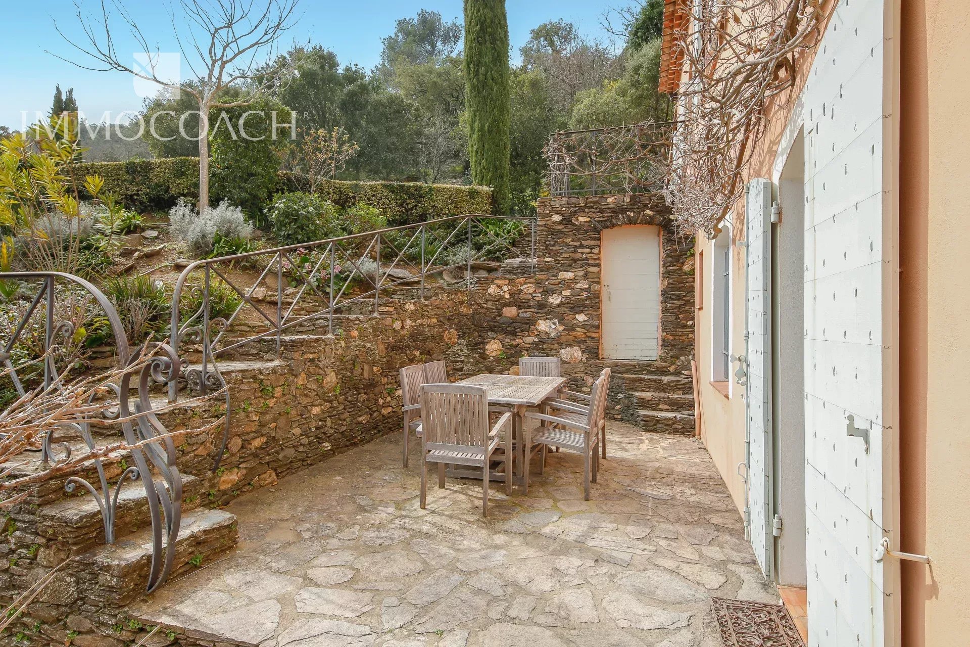 Stone patio with a wooden table and six chairs, surrounded by stone walls and garden, with a door and shutters on the right.