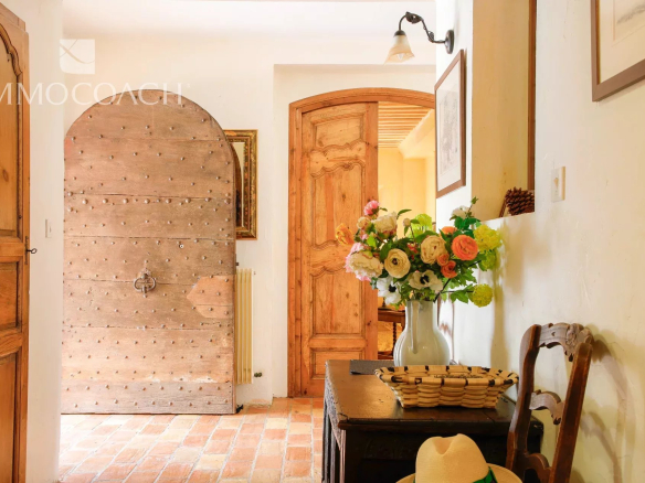 Rustic hallway with a large arched, studded wooden door to the left, and a bouquet of pink, white, and yellow flowers in a blue vase on a dark wooden table.