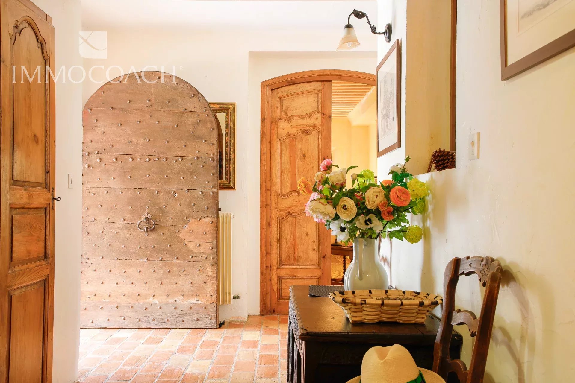 Rustic hallway with a large arched, studded wooden door to the left, and a bouquet of pink, white, and yellow flowers in a blue vase on a dark wooden table.
