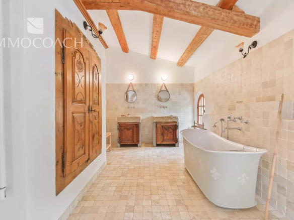 Rustic bathroom with a freestanding white tub, two stone sinks, exposed wooden ceiling beams, and beige tiled walls. An arched window brings in light.