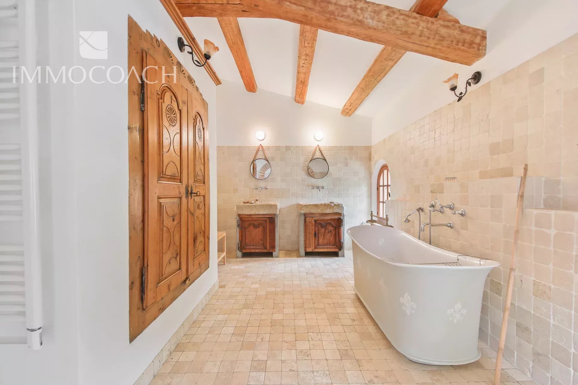 Rustic bathroom with a freestanding white tub, two stone sinks, exposed wooden ceiling beams, and beige tiled walls. An arched window brings in light.