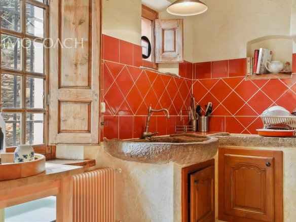 Rustic kitchen with red tile backsplash, stone sink, and wooden cabinets near a window. Pot rack and dish rack on the counter.