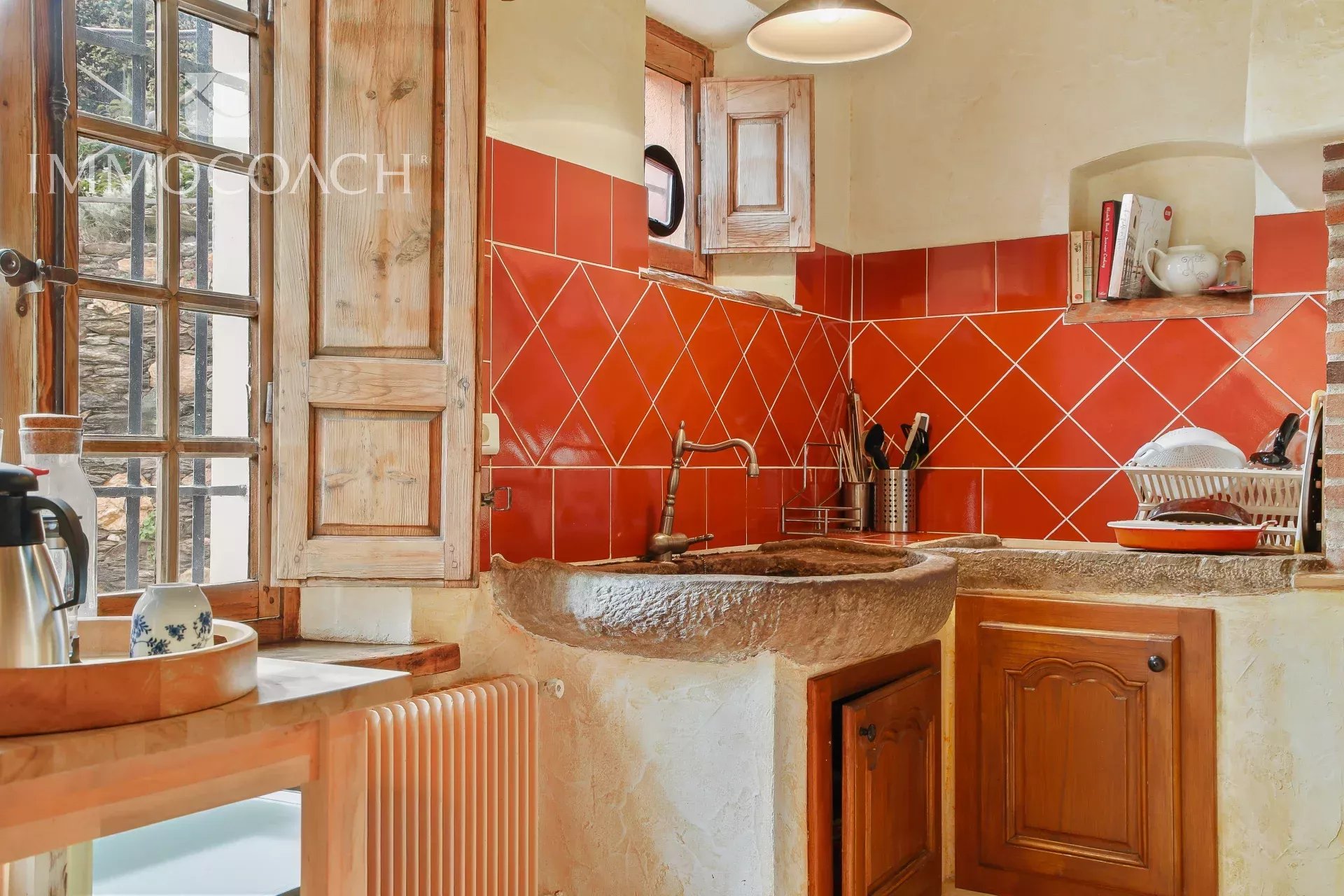 Rustic kitchen with red tile backsplash, stone sink, and wooden cabinets near a window. Pot rack and dish rack on the counter.