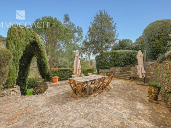 Stone patio with a wooden outdoor dining table and chairs, beige umbrellas, and trimmed hedges under a clear blue sky