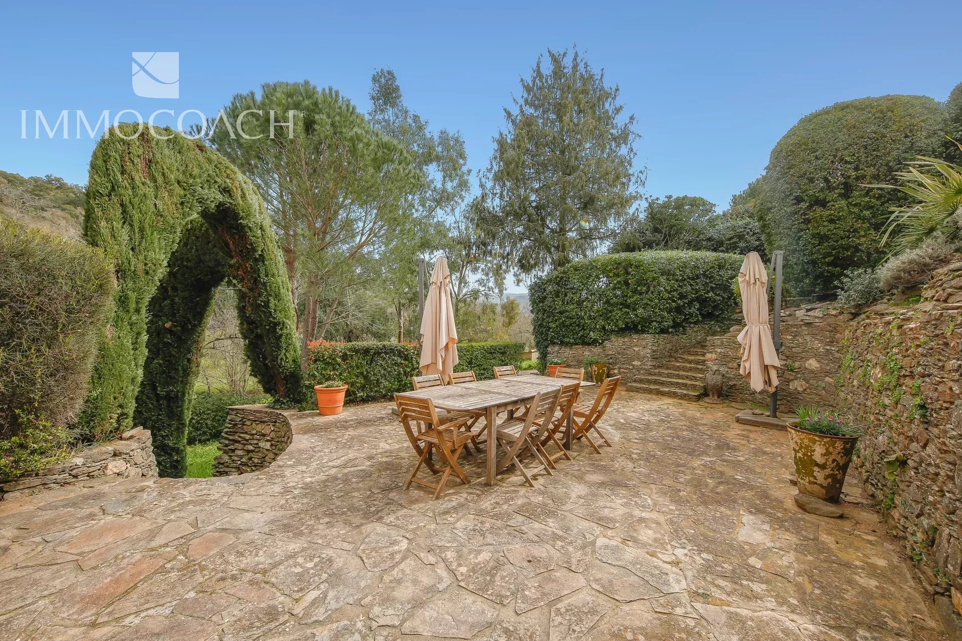 Stone patio with a wooden outdoor dining table and chairs, beige umbrellas, and trimmed hedges under a clear blue sky