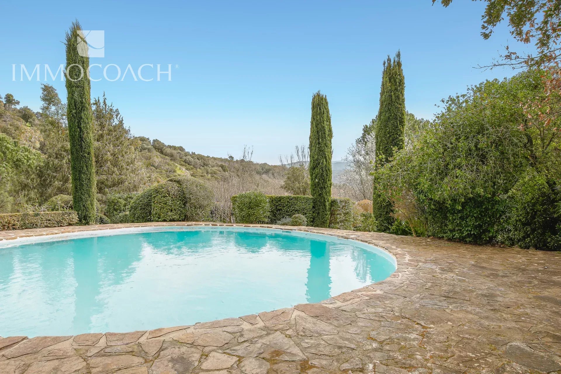 Kidney-shaped pool with turquoise water, bordered by a stone patio and tall cypress trees against a hilly landscape under a clear blue sky.