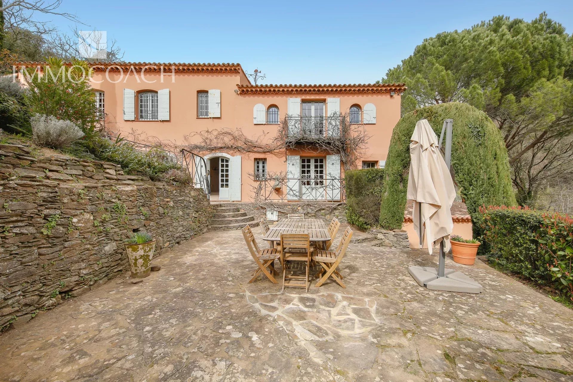 Mediterranean peach-colored villa with white shutters and a stone courtyard with a wooden dining set.