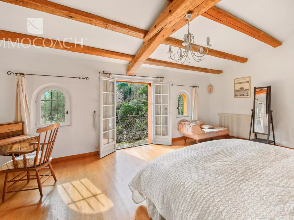 Bright bedroom with exposed wooden ceiling beams, French doors to a garden, and a white quilted bedspread in warm light