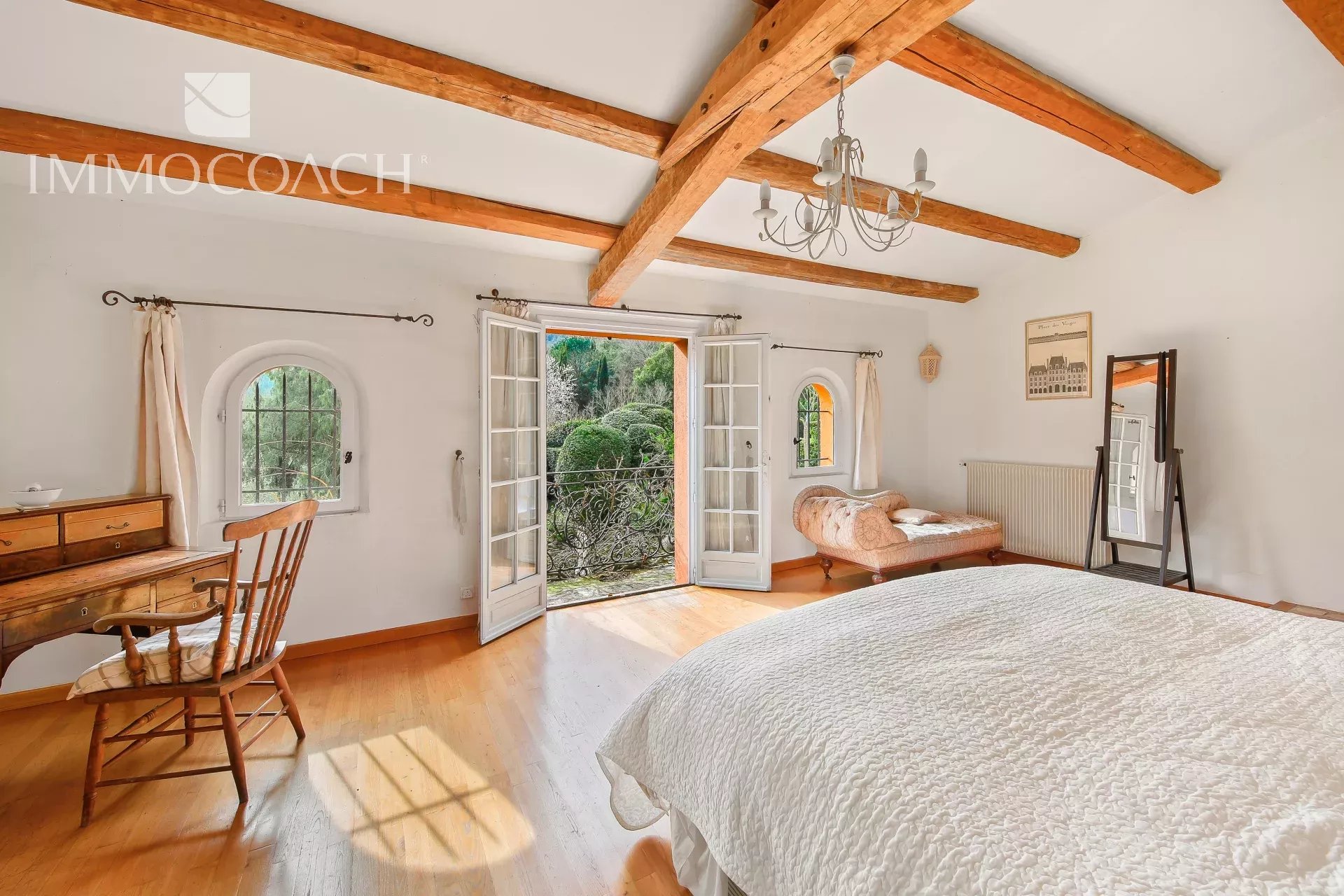 Bright bedroom with exposed wooden ceiling beams, French doors to a garden, and a white quilted bedspread in warm light
