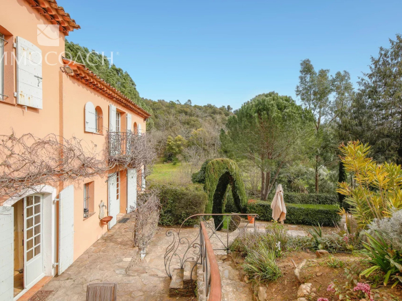 Peach-colored Mediterranean-style villa with white shutters and a small balcony along a stone patio, overlooking a garden.