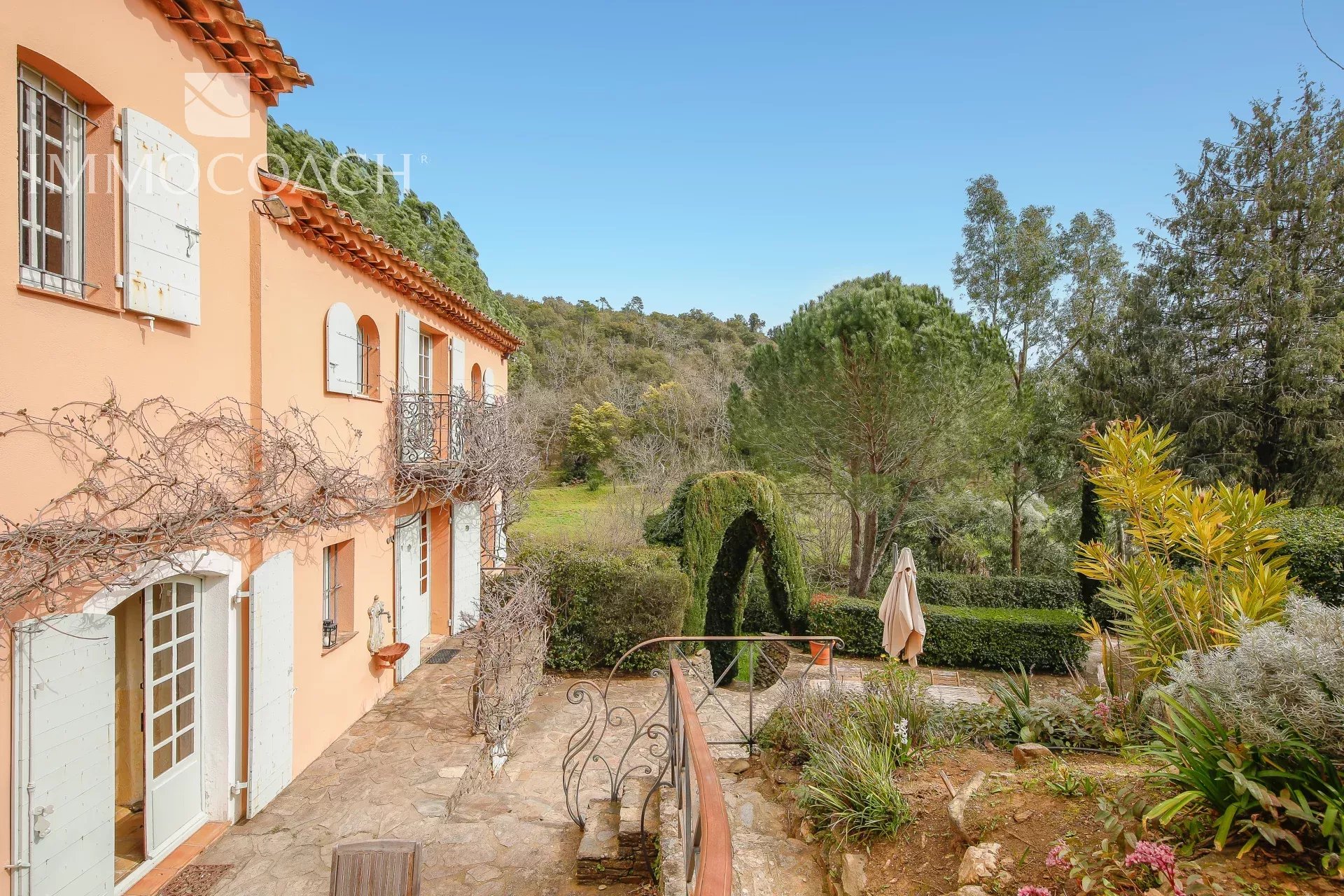 Peach-colored Mediterranean-style villa with white shutters and a small balcony along a stone patio, overlooking a garden.