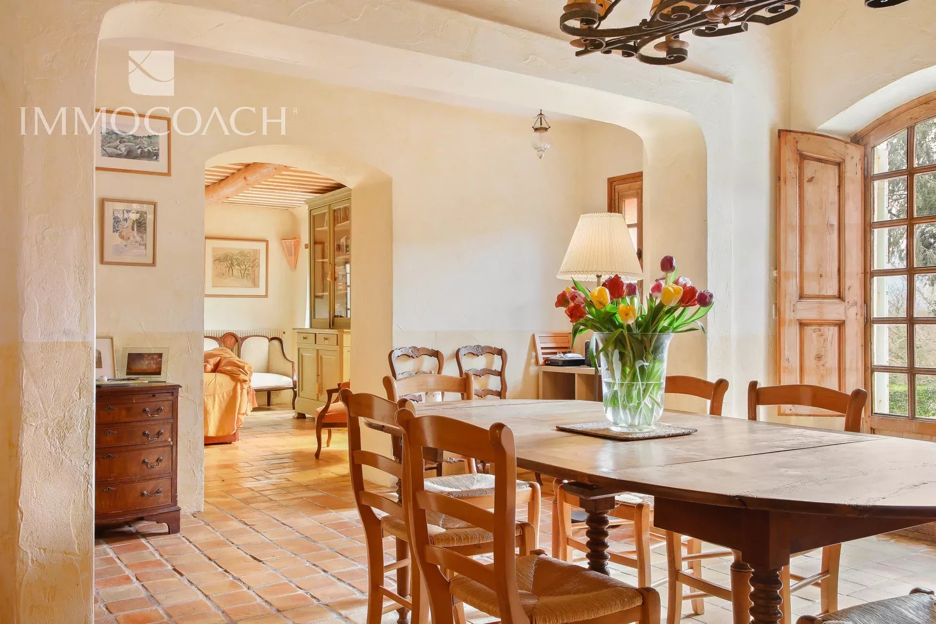 Cozy rustic dining room with a large wooden table, vase of tulips, and warm natural light from a window with shutters.