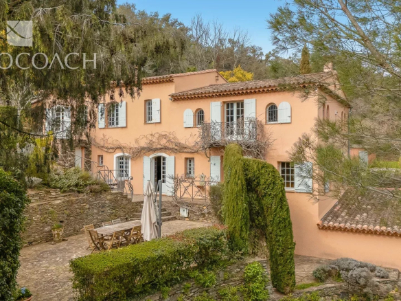 Pink Mediterranean villa with white shutters, iron balcony, and a stone courtyard framed by trees and greenery.