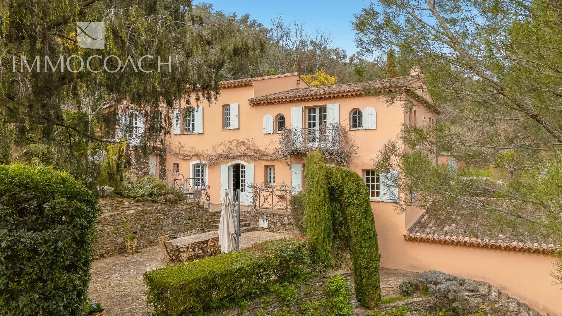 Pink Mediterranean villa with white shutters, iron balcony, and a stone courtyard framed by trees and greenery.