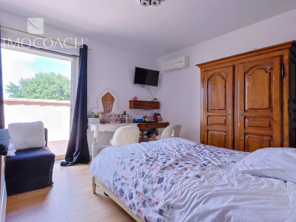 Cozy bedroom with floral duvet, white furniture, and a large wooden armoire; balcony door open to sunny outdoors.