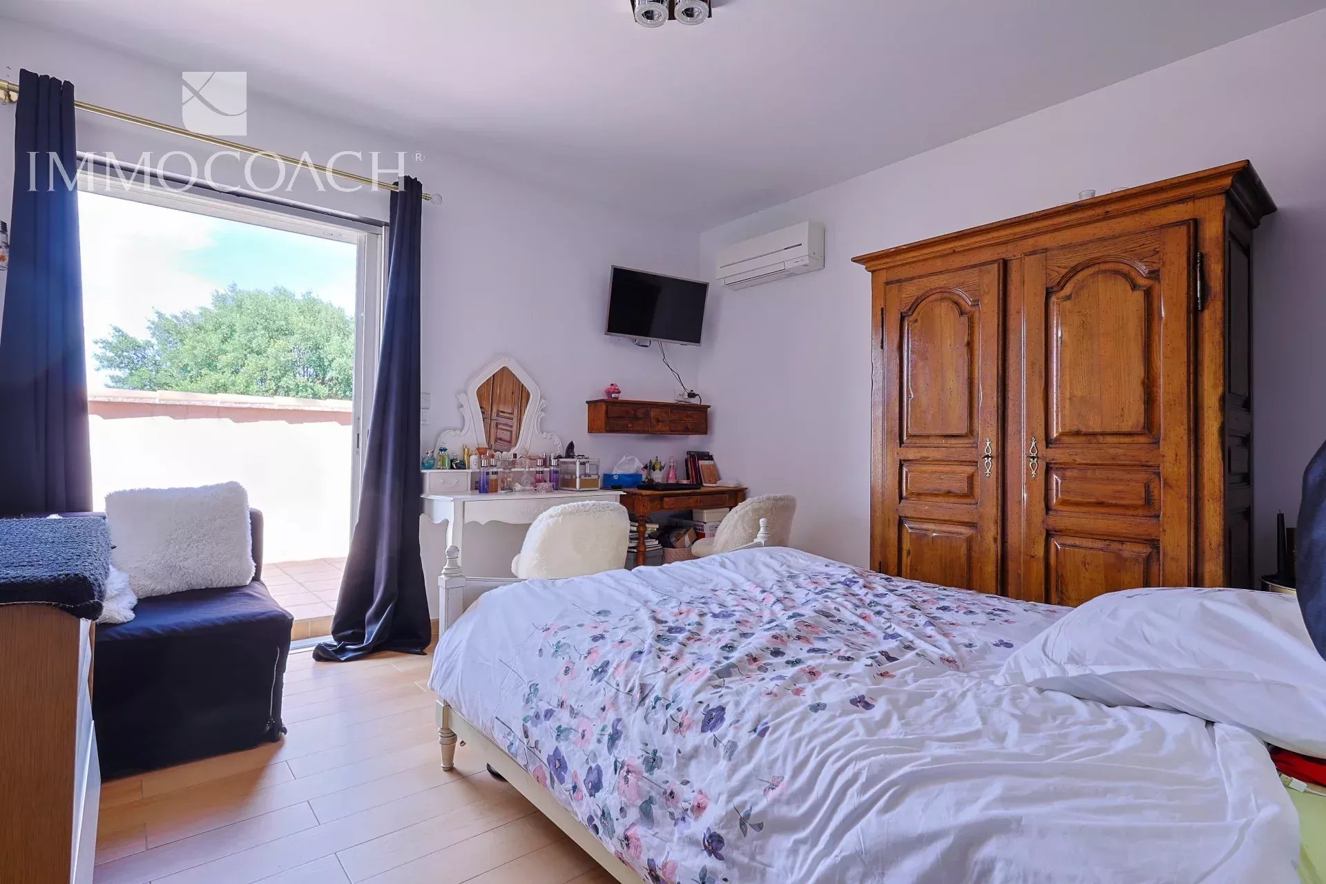 Cozy bedroom with floral duvet, white furniture, and a large wooden wardrobe; balcony door open to sunny outdoors.