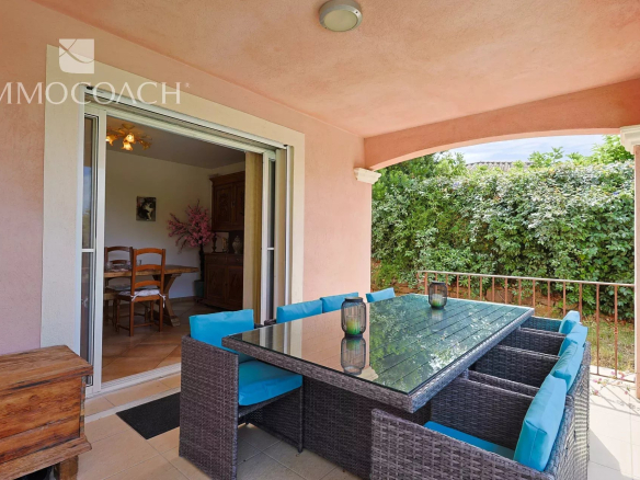 Covered patio with a glass-topped wicker dining table and blue-cushioned chairs, opening to a dining room; green hedge beyond.