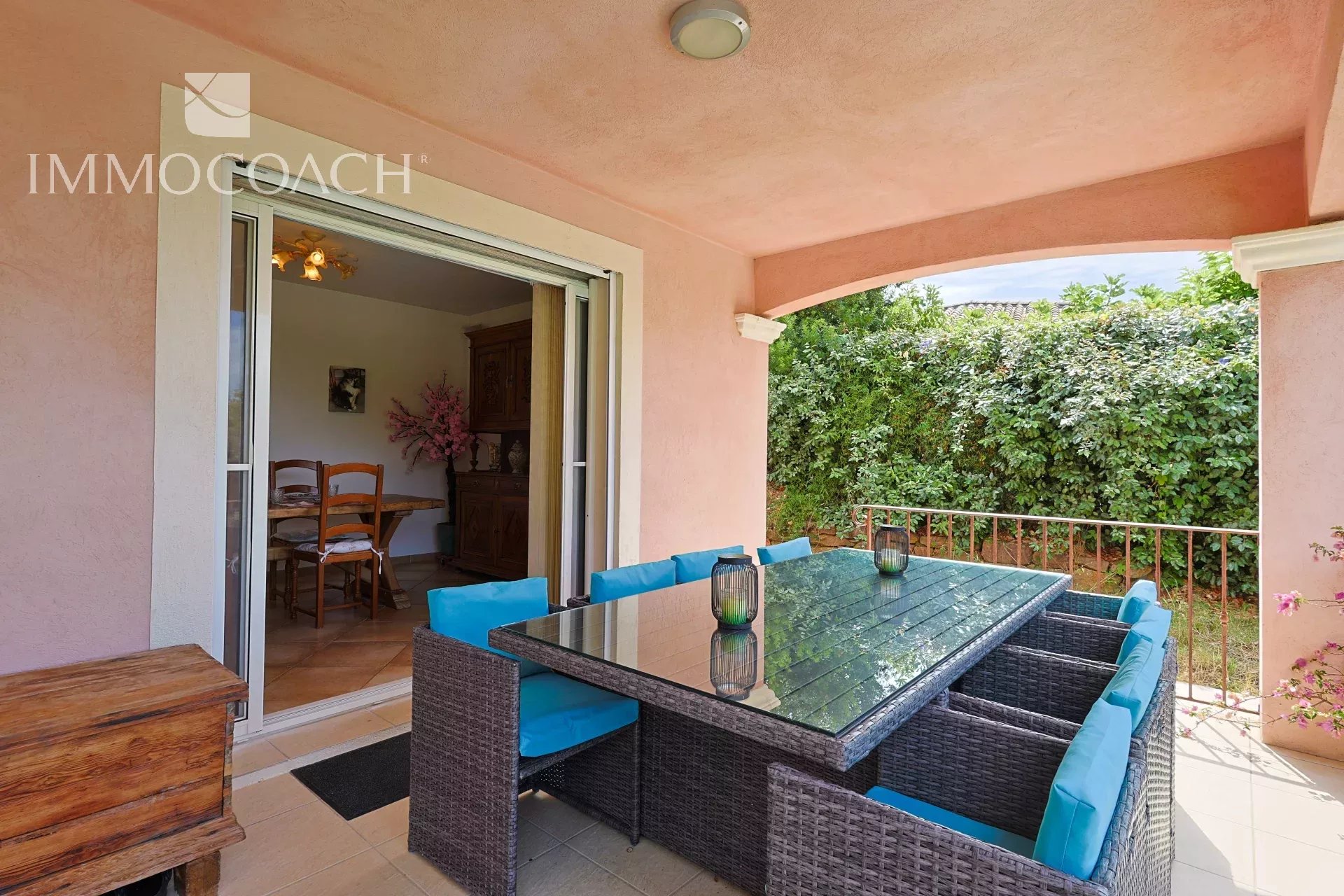 Covered patio with a glass-topped wicker dining table and blue-cushioned chairs, opening to a dining room; green hedge beyond.