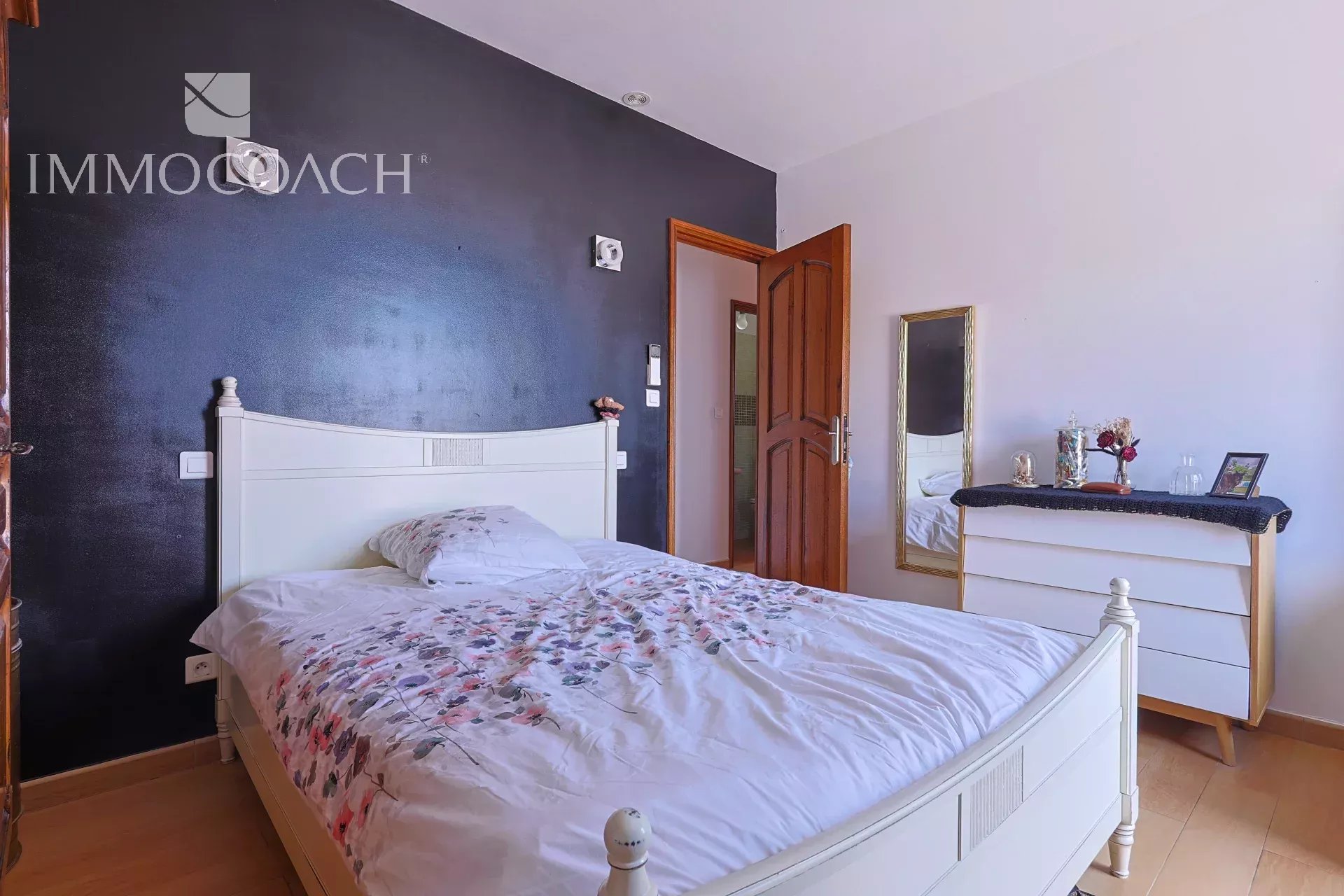 White wooden bed with floral duvet in a bedroom with a dark blue accent wall.