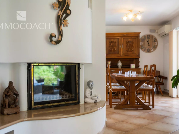 Cozy open-plan living and dining area with a curved white fireplace, wooden dining table and chairs, a large round wall clock, and a leafy plant by the glass door.