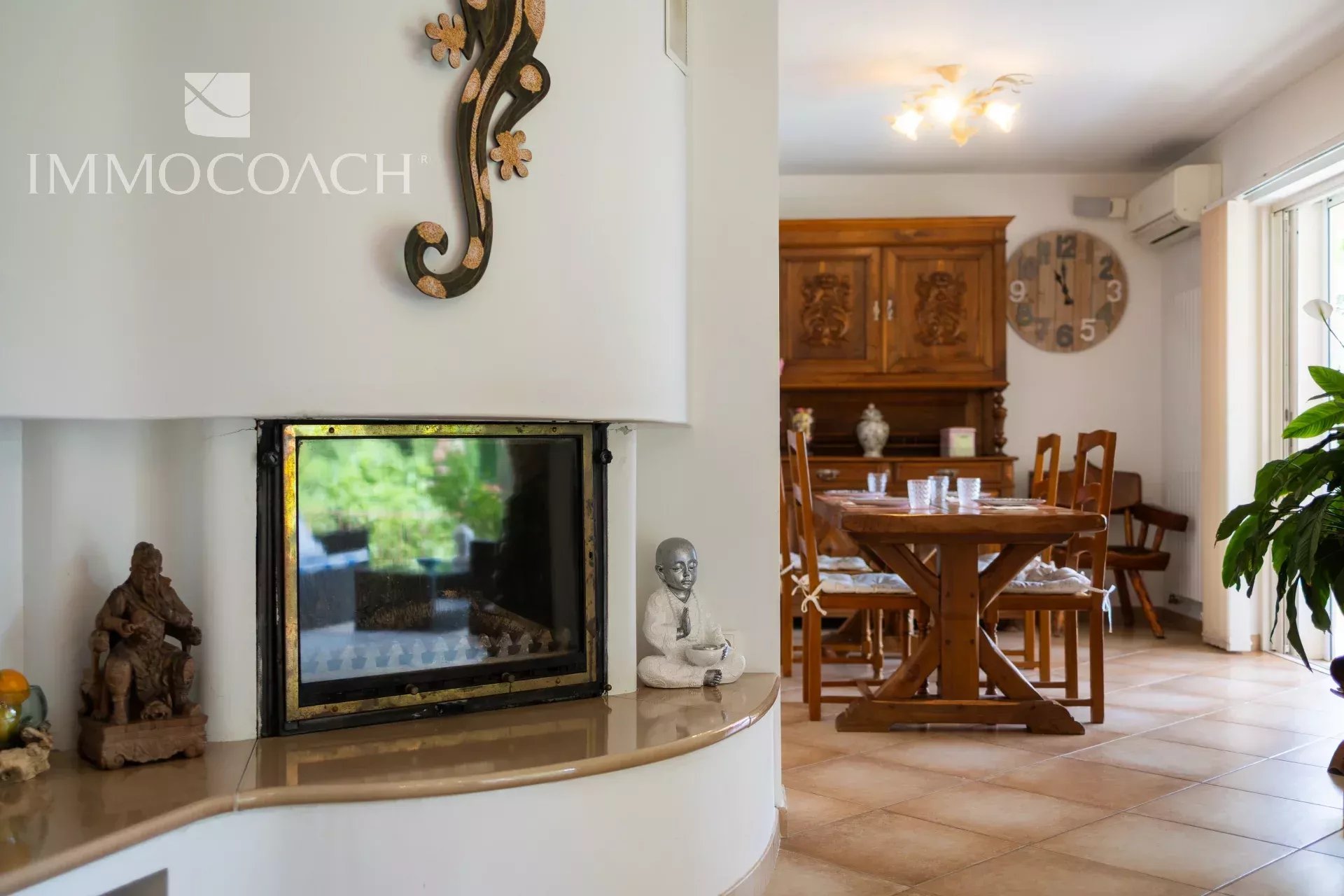 Cozy open-plan living and dining area with a curved white fireplace, wooden dining table and chairs, a large round wall clock, and a leafy plant by the glass door.