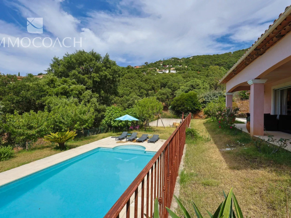 Backyard with a rectangular pool, six lounge chairs and a blue umbrella, surrounded by green trees and a pink patio house on the right.