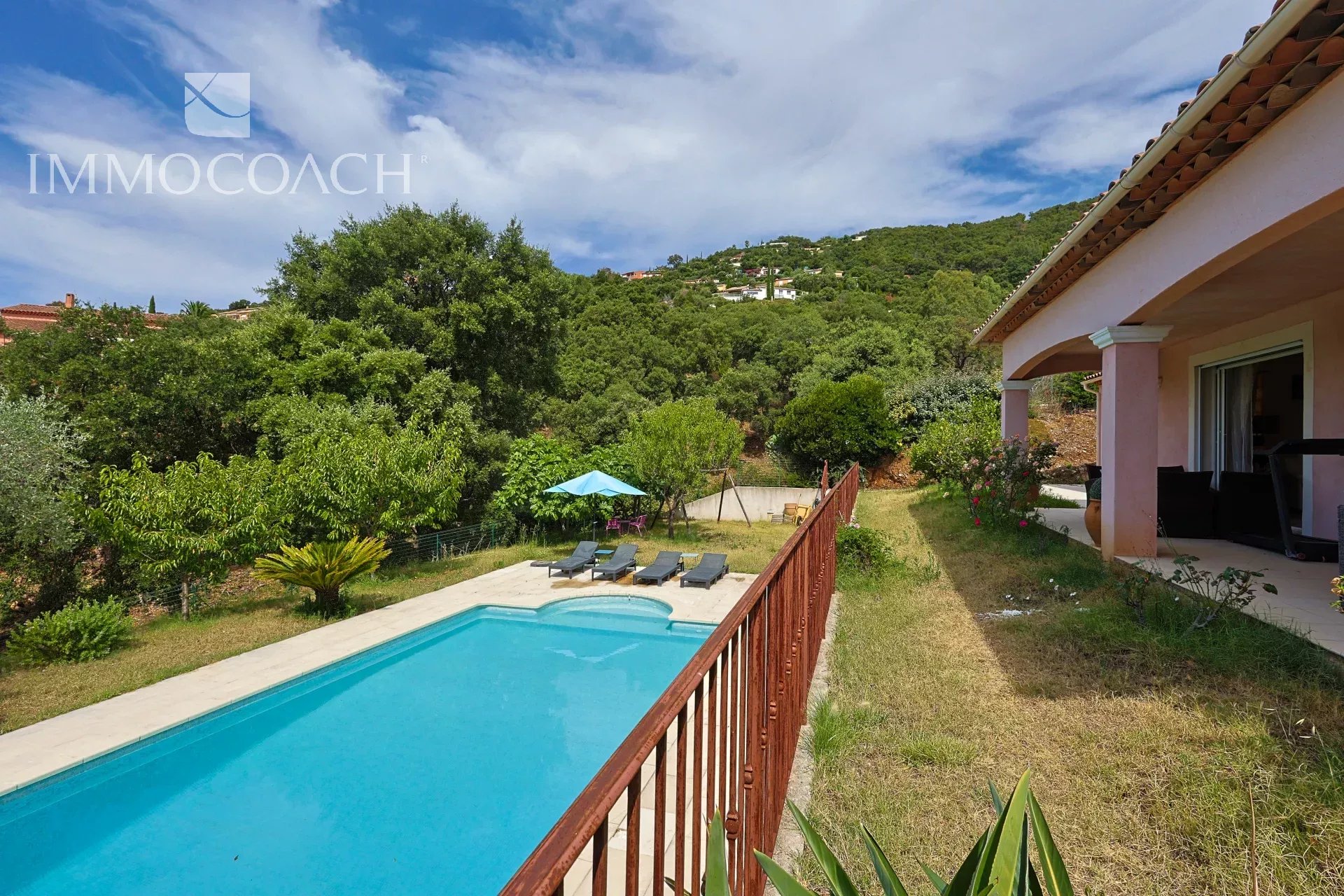 Backyard with a rectangular pool, six lounge chairs and a blue umbrella, surrounded by green trees and a pink patio house on the right.