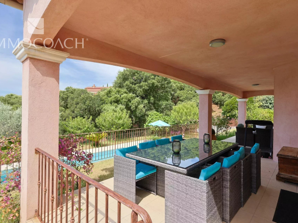 Covered outdoor patio with a glass-top wicker dining table and turquoise cushions, overlooking a pool and lush garden.
