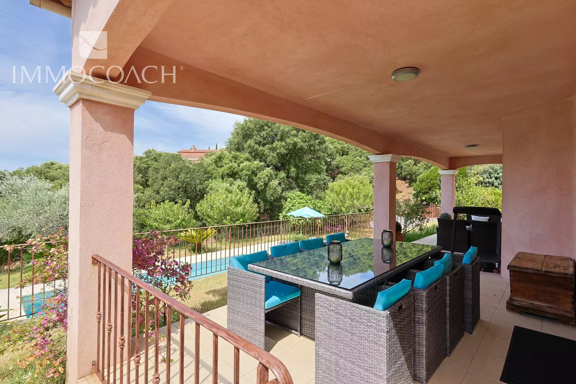Covered outdoor patio with a glass-top wicker dining table and turquoise cushions, overlooking a pool and lush garden.