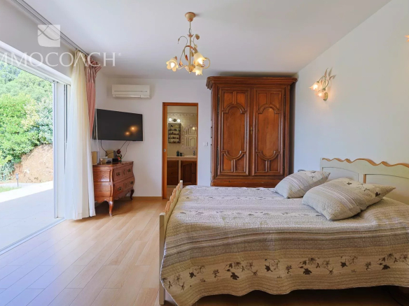 Cozy bedroom with a large wooden armoire, a double bed with quilted bedding, and a sliding glass door opening to a green garden outside.