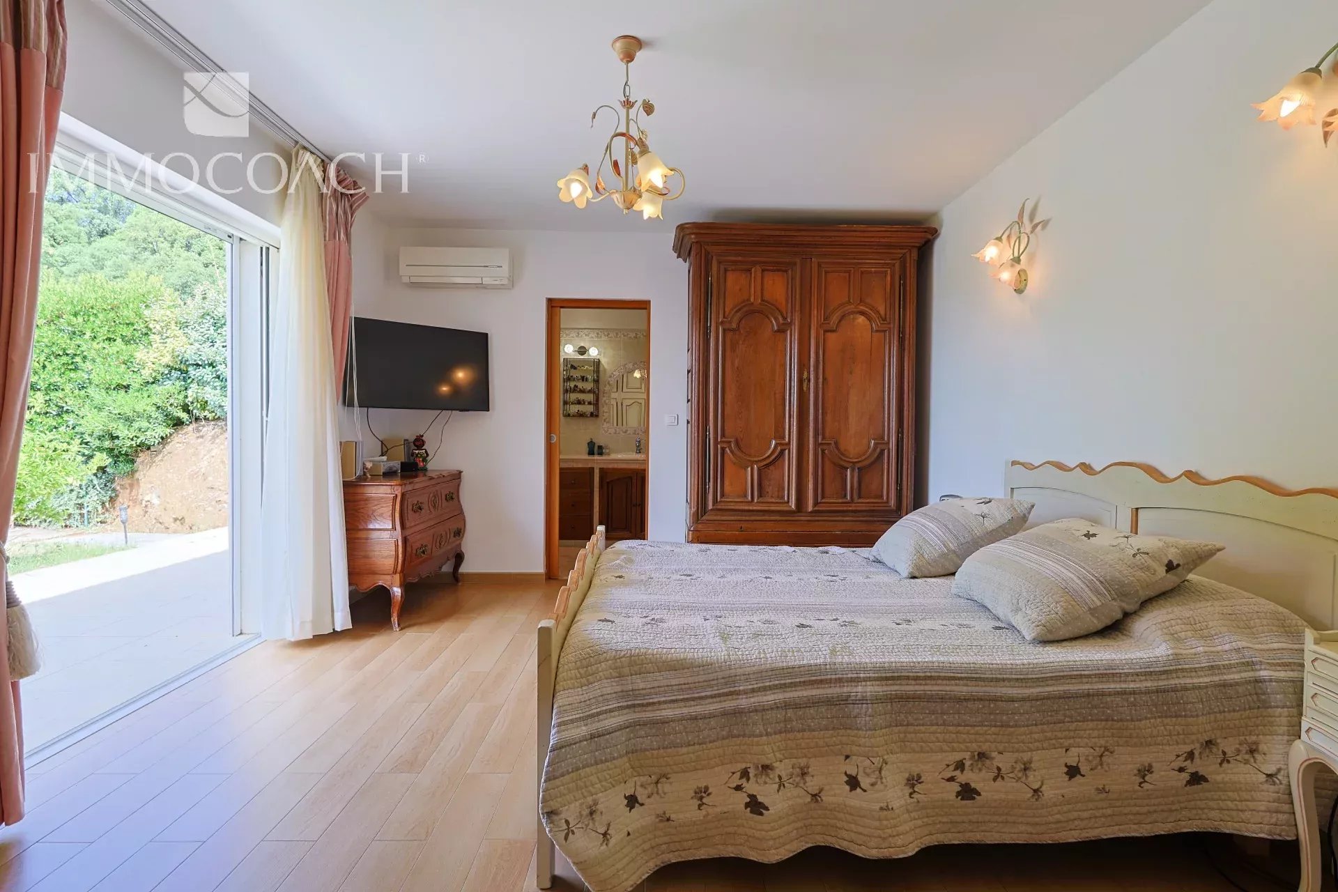 Cozy bedroom with a large wooden wardrobe, a double bed with quilted bedding, and a sliding glass door opening to a green garden outside.