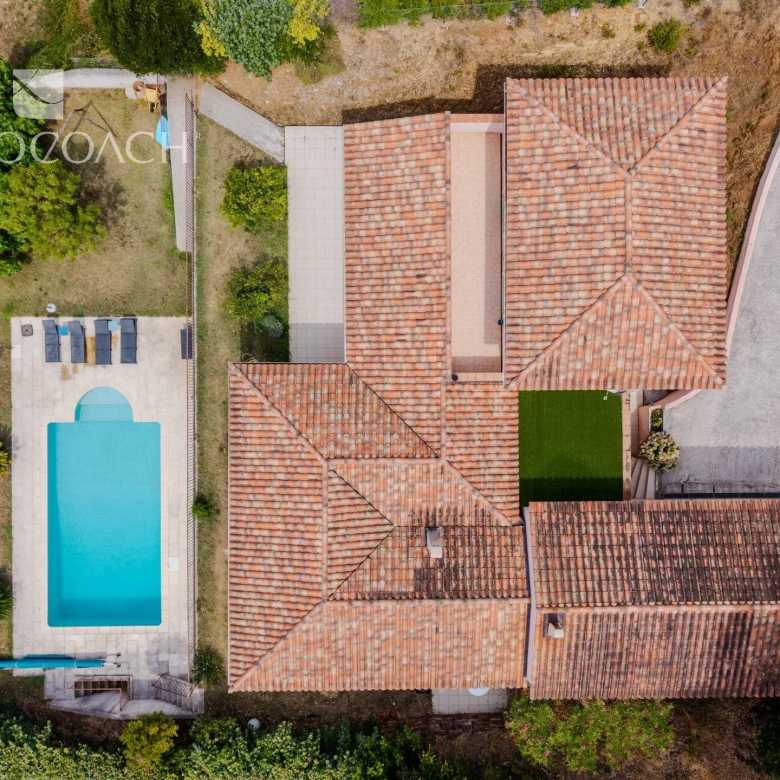 Aerial view of a Mediterranean-style villa with a terracotta-tiled roof, a rectangular blue pool, and a lounge area on a light stone patio to the left.