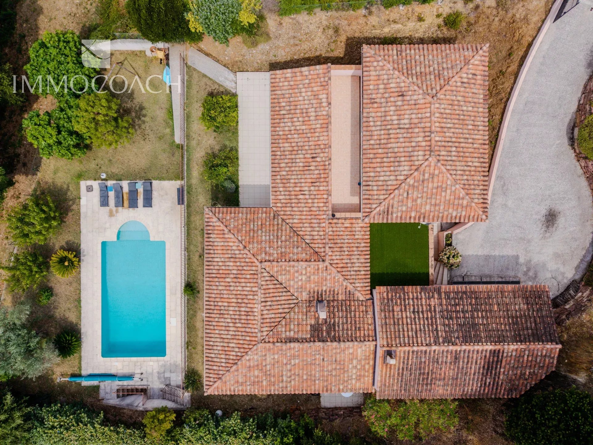Aerial view of a Mediterranean-style villa with a terracotta-tiled roof, a rectangular blue pool, and a lounge area on a light stone patio to the left.