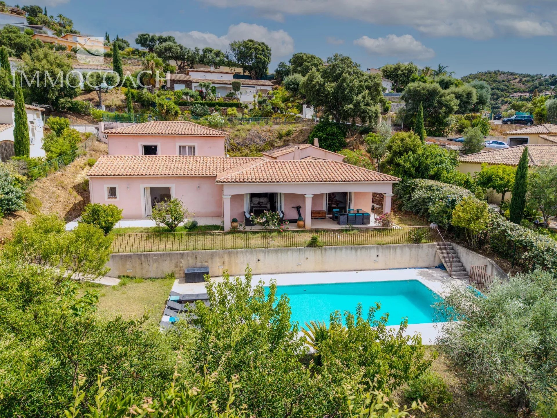 Pink Mediterranean-style villa with tiled roof and a rectangular blue pool in the yard, surrounded by trees and hills.