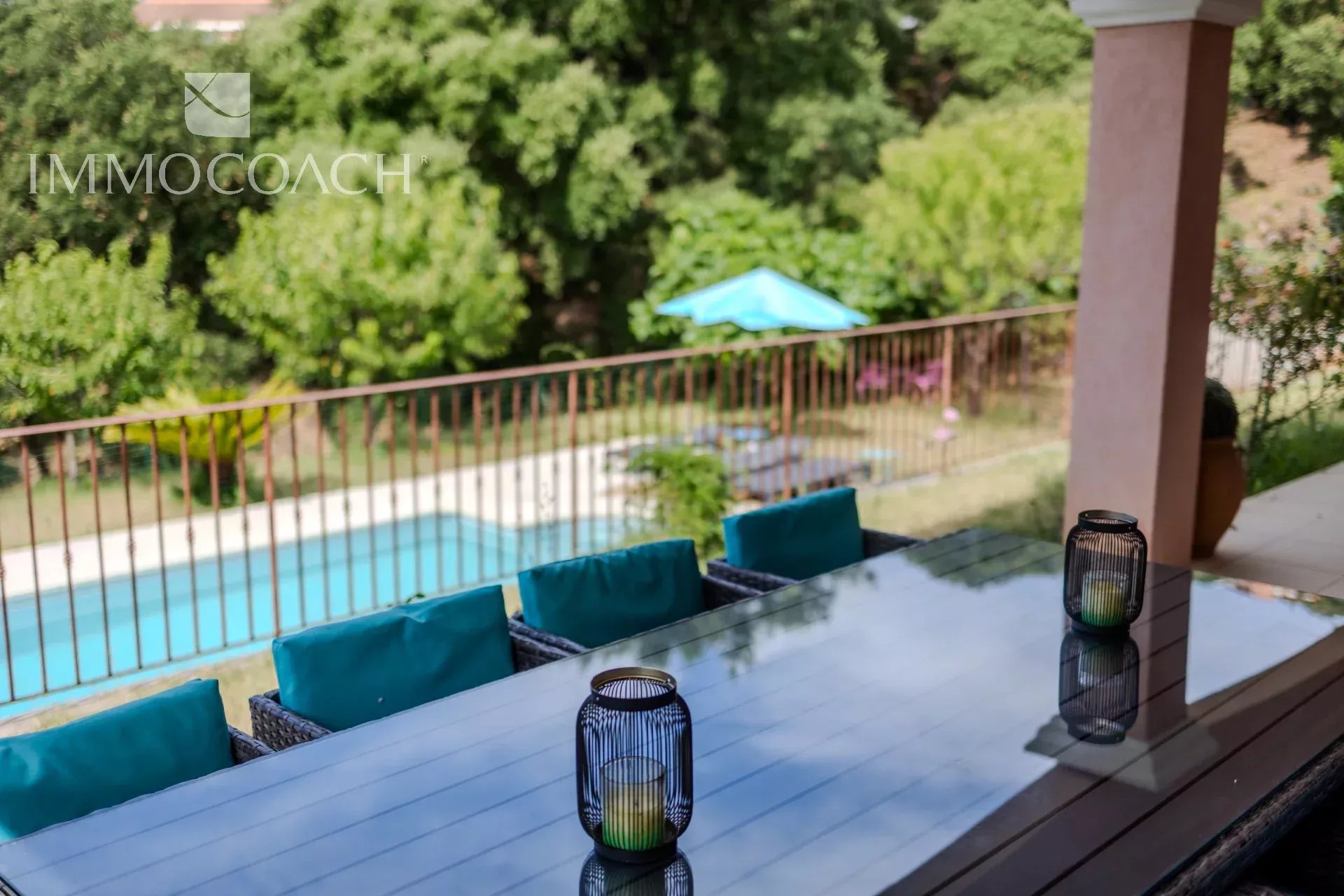 Patio scene with glass table, teal cushions, blue lanterns, and a pool beyond; ImmoCoach watermark visible.