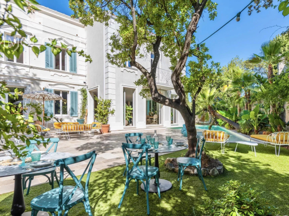 Sunny courtyard with a white building, turquoise chairs around tables, yellow cushioned benches along a pool, and lush trees with string lights overhead.