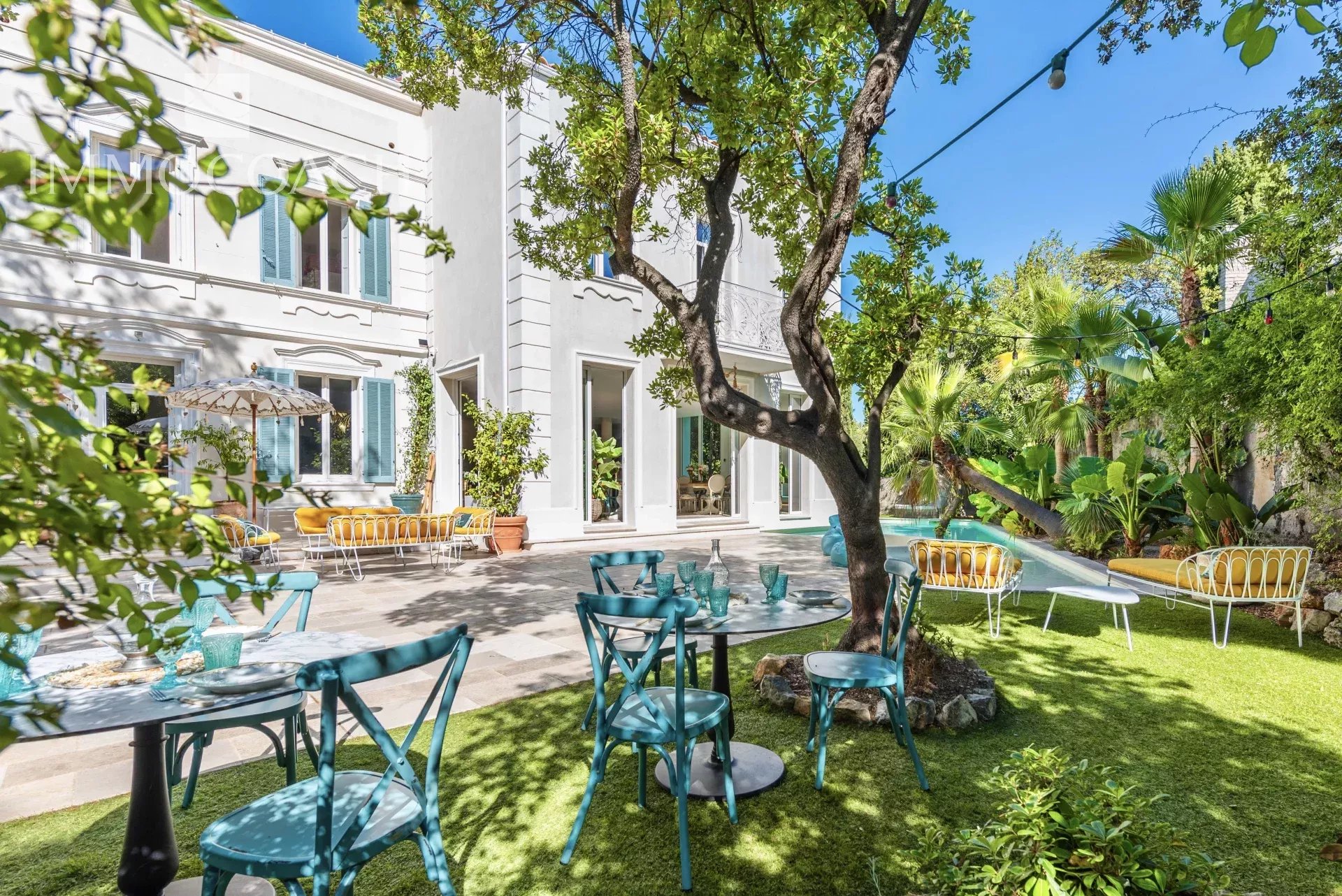 Sunny courtyard with a white building, turquoise chairs around tables, yellow cushioned benches along a pool, and lush trees with string lights overhead.