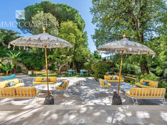 Sunny outdoor lounge area with yellow-cushioned white metal sofas under decorative umbrellas on a stone patio, surrounded by green trees and plants.